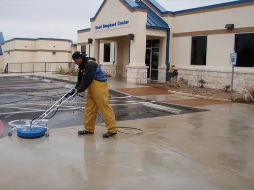 Technician in yellow waders running a rotary surface cleaner across a wet commercial plaza